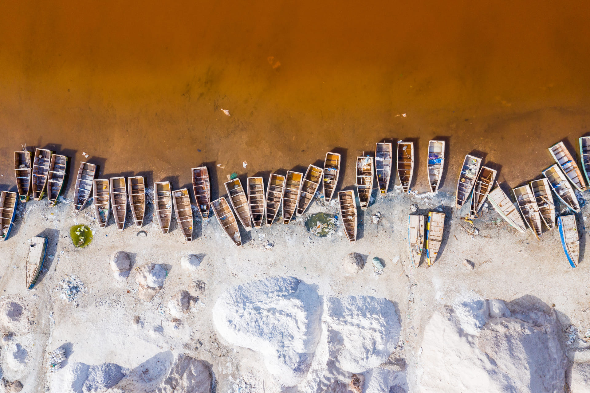 Aerial view of wooden boats along a rust-colored lakeshore with white salt mounds