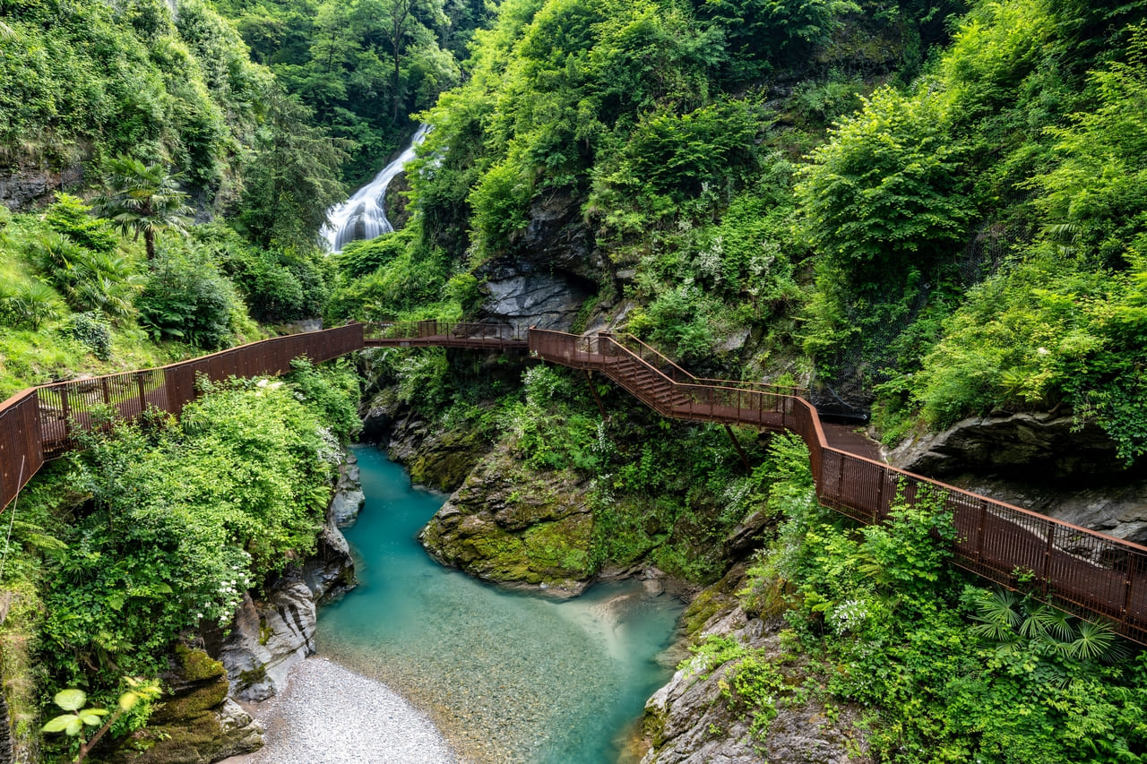 A wooden walkway winds along a lush gorge with turquoise waters and a cascading waterfall in Bellano, Italy